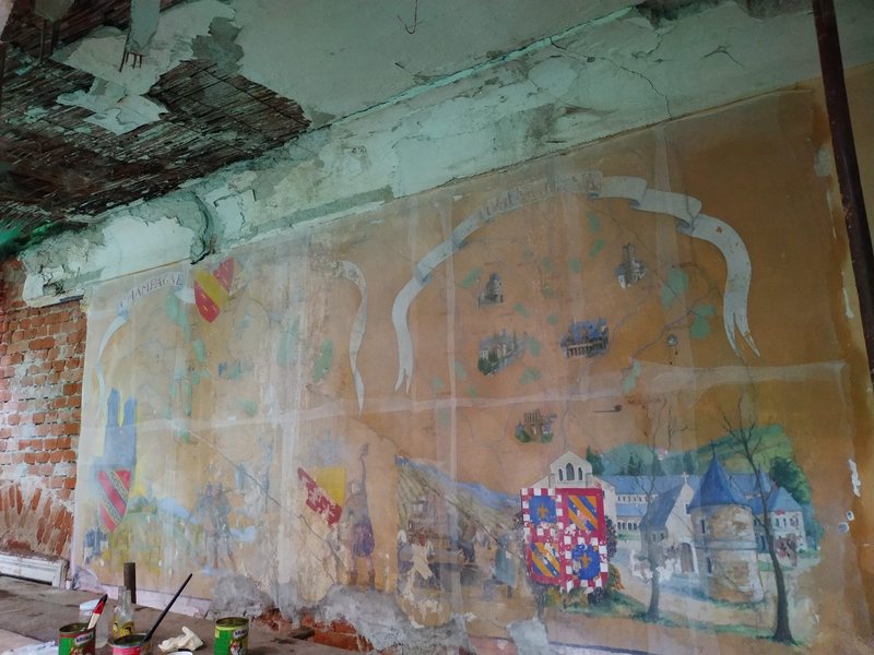 Nástěnná malba s krajinou, budovami a erby na stěně zámku pod poškozeným stropem, před sejmutím
